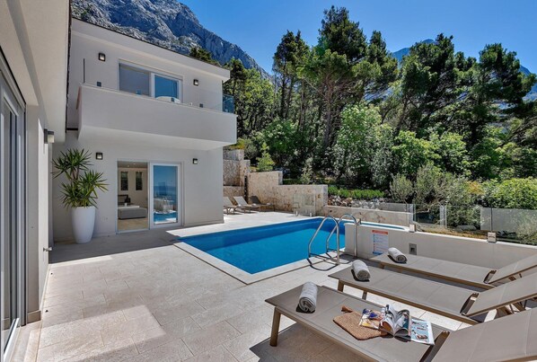 A modern two-story building with a swimming pool, outdoor seating, and a mountainous backdrop.