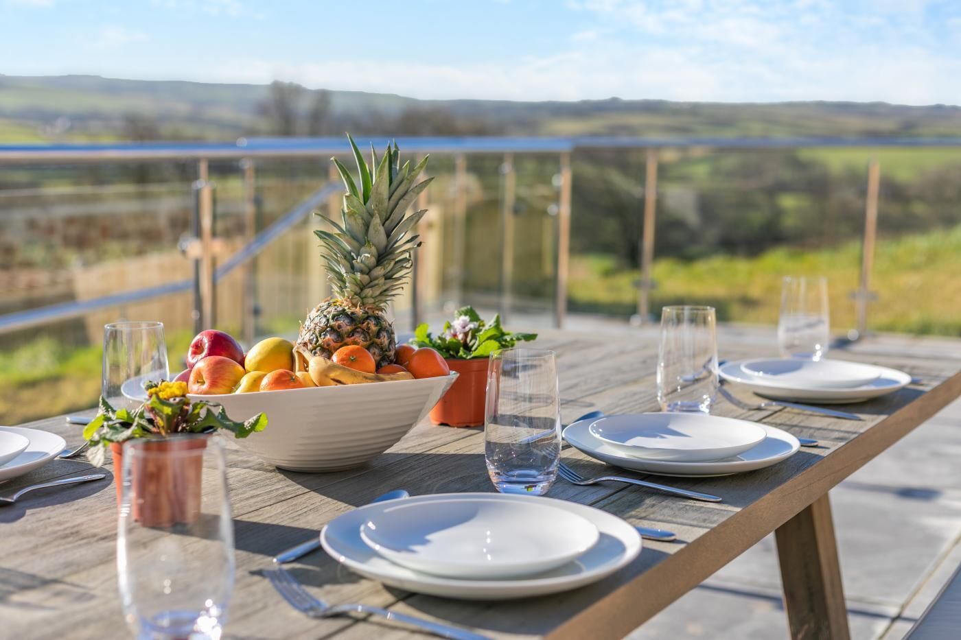 A wooden table set for a meal with a bowl of fruit, glasses, and plates, overlooking a scenic landscape.