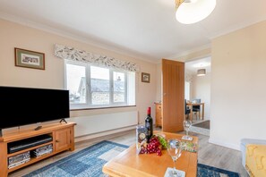 Ein Wohnzimmer mit Fernseher, Holzmöbeln, einem Esstisch mit Flasche und Gläsern sowie einem Fenster mit Blick auf Häuser.