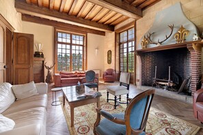 A spacious living room with a fireplace, wooden ceiling beams, and a large window. - Château du Perray – Princess Room with Park View and Shared Pool (Saint-Mars-du-Désert)