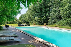 Une piscine entourée d’une végétation luxuriante, avec des chaises longues et une petite table à proximité.