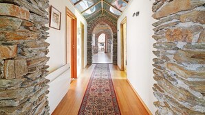 A stone-walled hallway with a wooden floor and a patterned rug, leading to a room with a skylight.