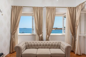 Ein Zimmer mit einem beigefarbenen Sofa, zwei Fenstern mit beigen Vorhängen und Meerblick.