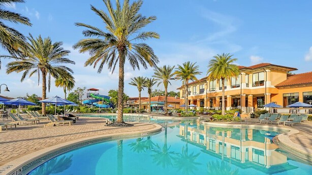 Una piscina rodeada de palmeras, sillas de descanso y edificios de estilo mediterráneo.