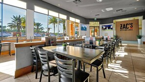 A spacious restaurant with high ceilings, large windows offering views of palm trees, and a bar area with a sign reading "ZAC'S Sports Bar & Grill".