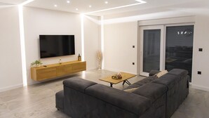 Ein modernes Wohnzimmer mit einem großen Flachbildfernseher, einem grauen Sofa, einem Holztisch und einer Schiebetür zum Balkon.