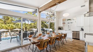 A modern kitchen with white cabinets, a stainless steel dishwasher, and a dining area with a wooden table and chairs. - Spacious Family Retreat w/ Pool, LSV, 7 Bikes, & Generous Outdoor Space w/BBQ (Dune Allen)