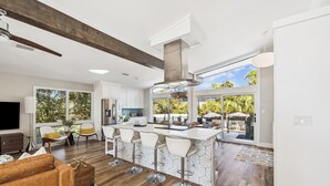 A modern kitchen with a central island, bar stools, and a large window overlooking a patio. - Spacious Family Retreat w/ Pool, LSV, 7 Bikes, & Generous Outdoor Space w/BBQ (Dune Allen)