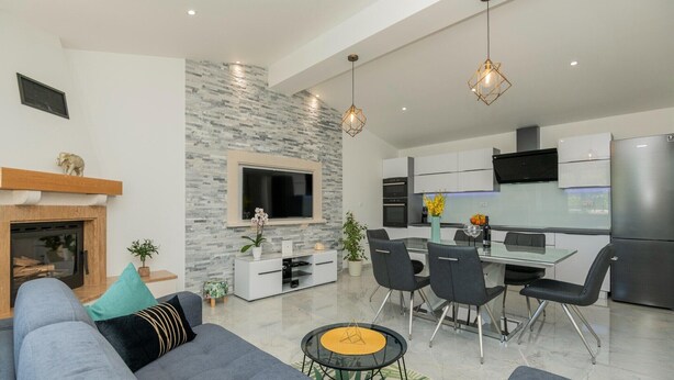 A modern living room with a fireplace, grey sofa, dining area with chairs, and a kitchen with appliances. - Tranquil Country Villa in Naklice (županija,)