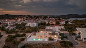 A coastal town at dusk with a prominent modern house featuring a swimming pool, surrounded by residential buildings and a body of water.