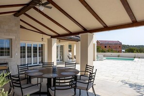 A covered patio with a round table and chairs, a pool, and a view of a house in the background.