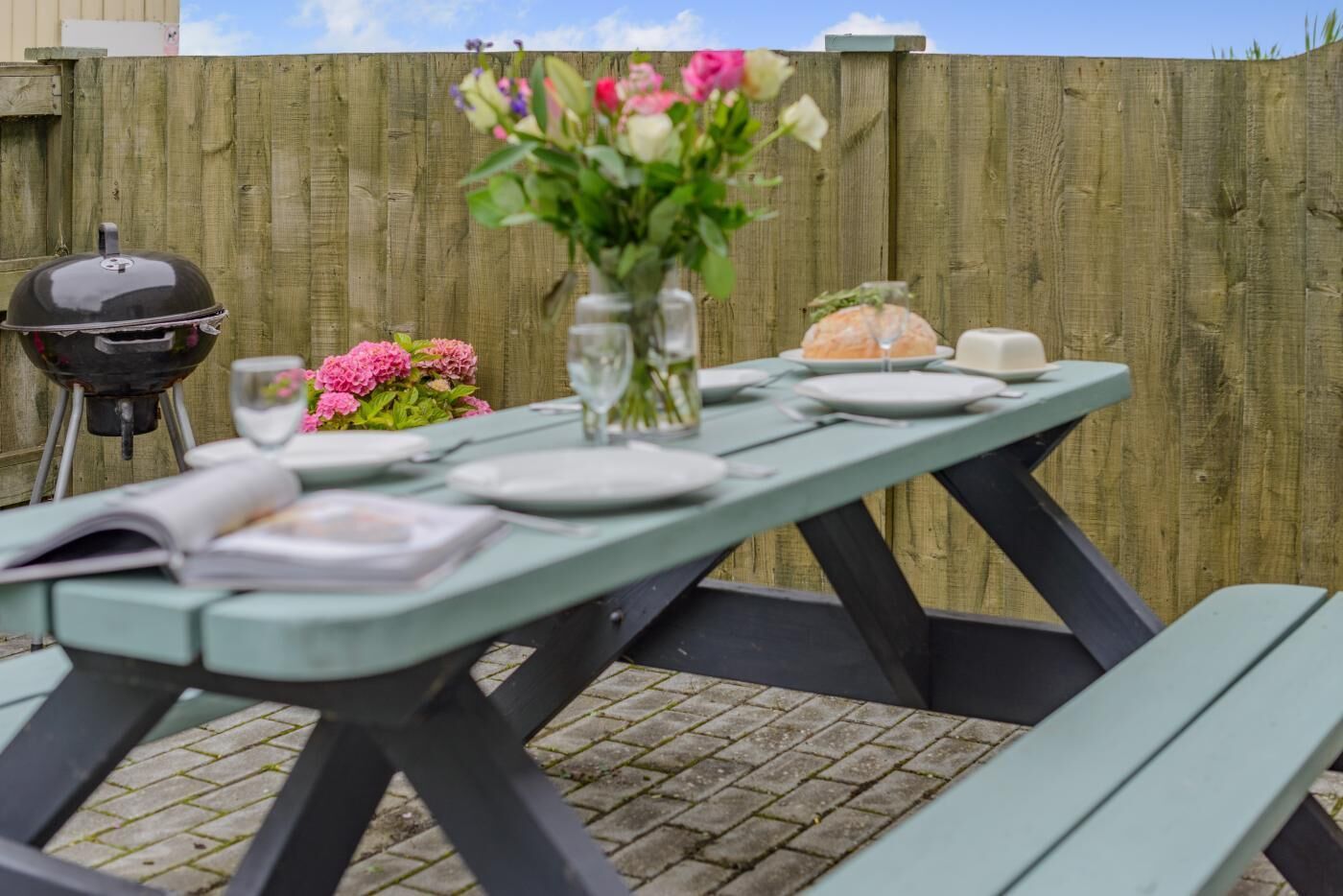 A picnic table set outdoors with a grill, a vase with flowers, and a book.