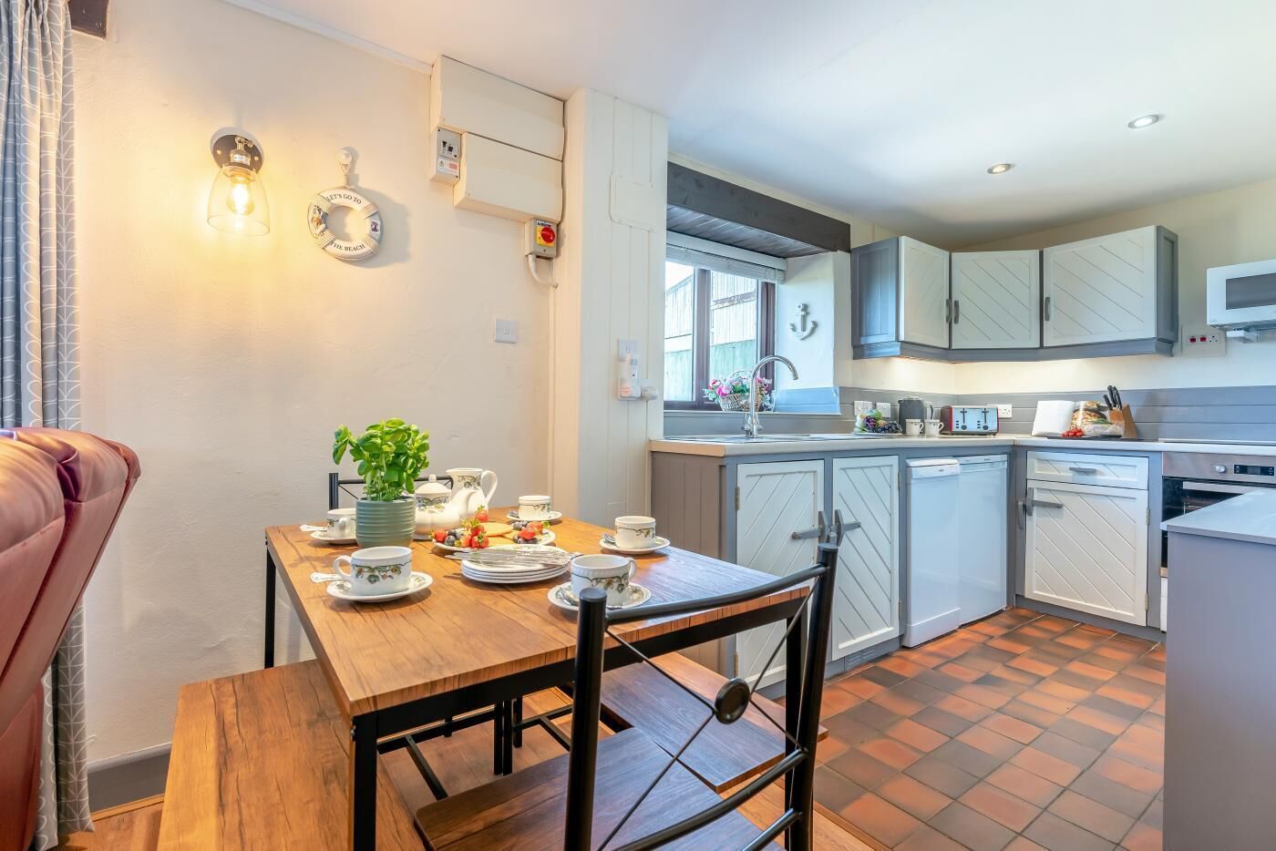 A modern kitchen with a wooden dining table set for tea, a potted plant, and a view of the adjacent dining area.