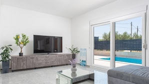 Ett modernt vardagsrum med en platt-TV, ett soffbord i glas, en grå soffa och en skjutdörr i glas som leder ut till en pool.