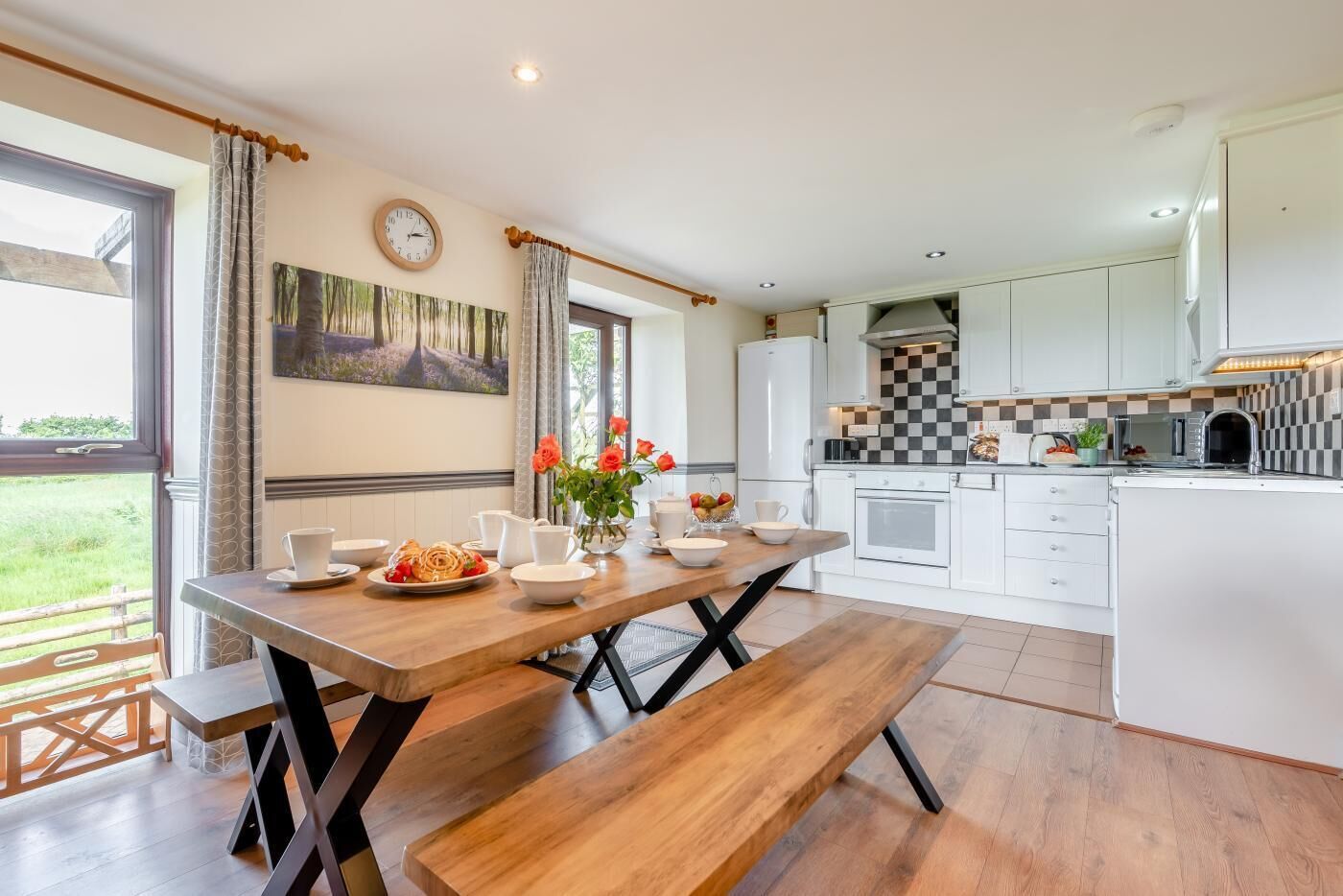 Una cocina moderna con armarios blancos, una mesa de comedor de madera y un banco. Hay un reloj en la pared y un cuadro sobre el lavadero.