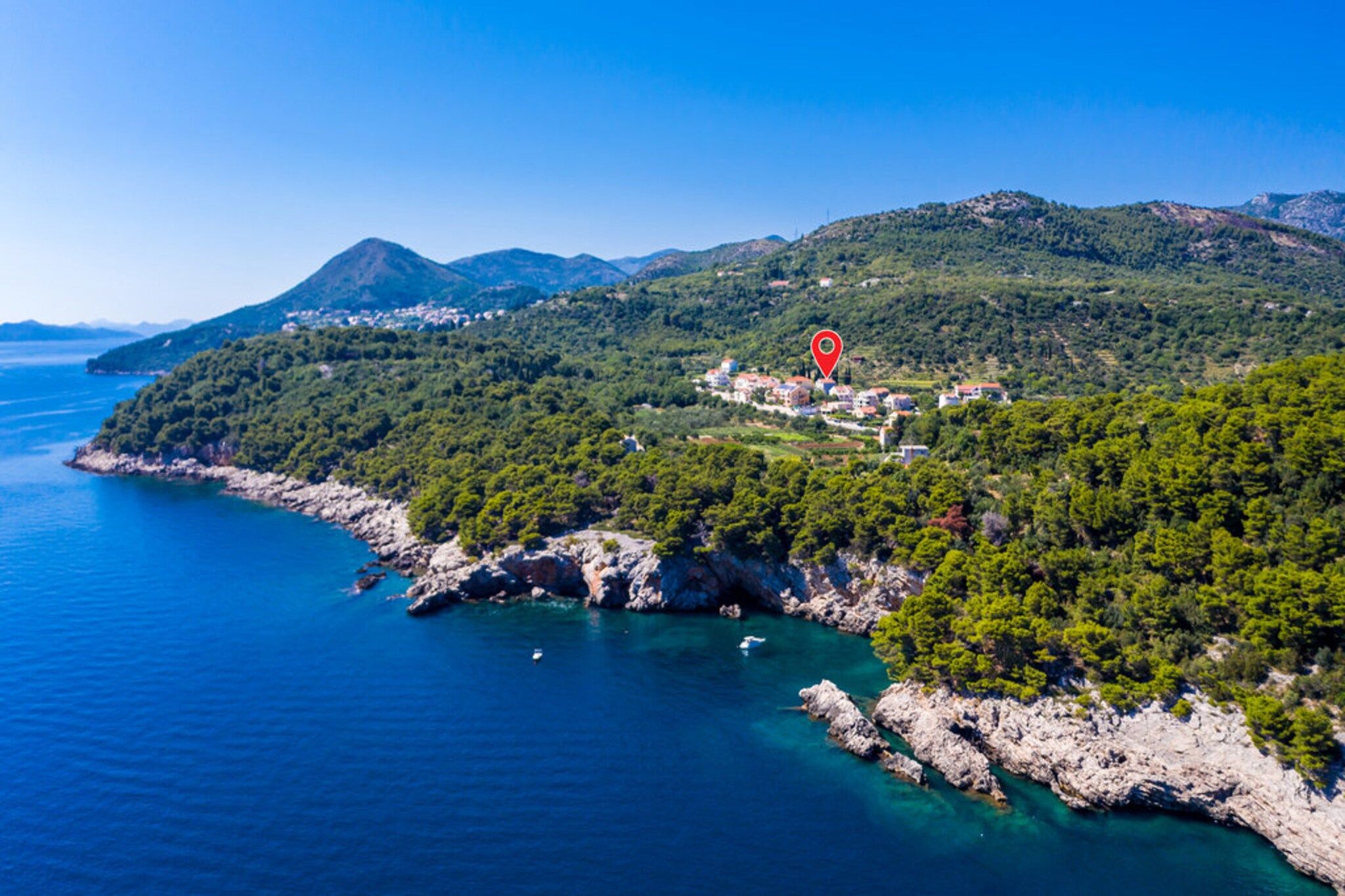 A coastal landscape with a red location marker, rocky shoreline, clear blue sea, and a village on a hillside.