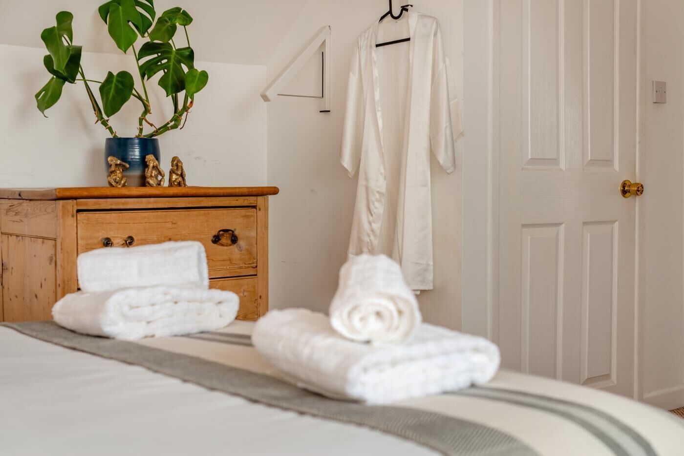 A neatly made bed with rolled towels, a wooden dresser with a plant, and a white robe hanging on a hook.