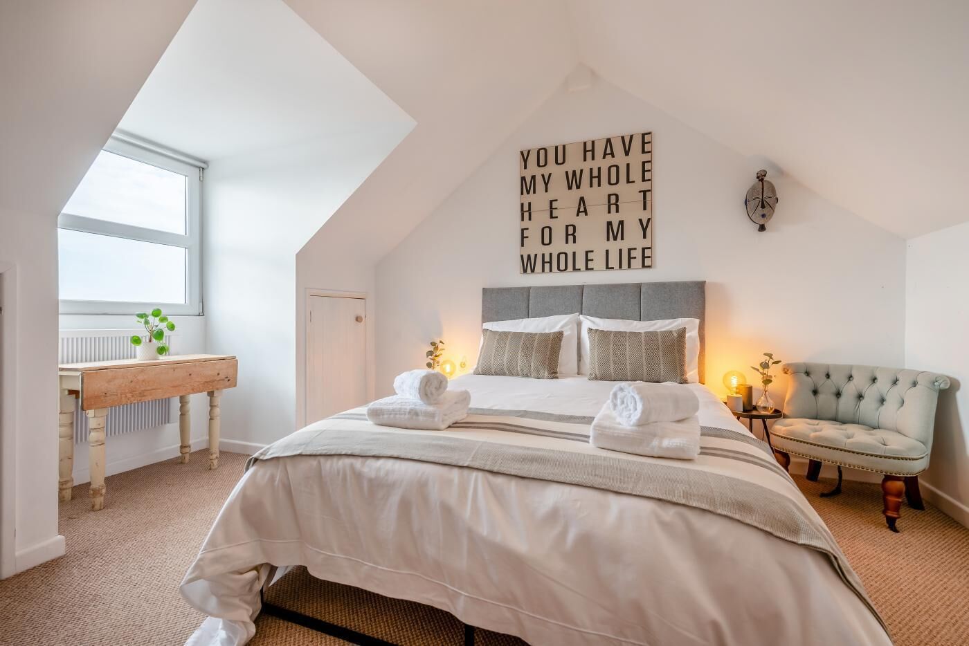 A bedroom with a bed, a bedside table, a chair, a wall with a message, and a window.