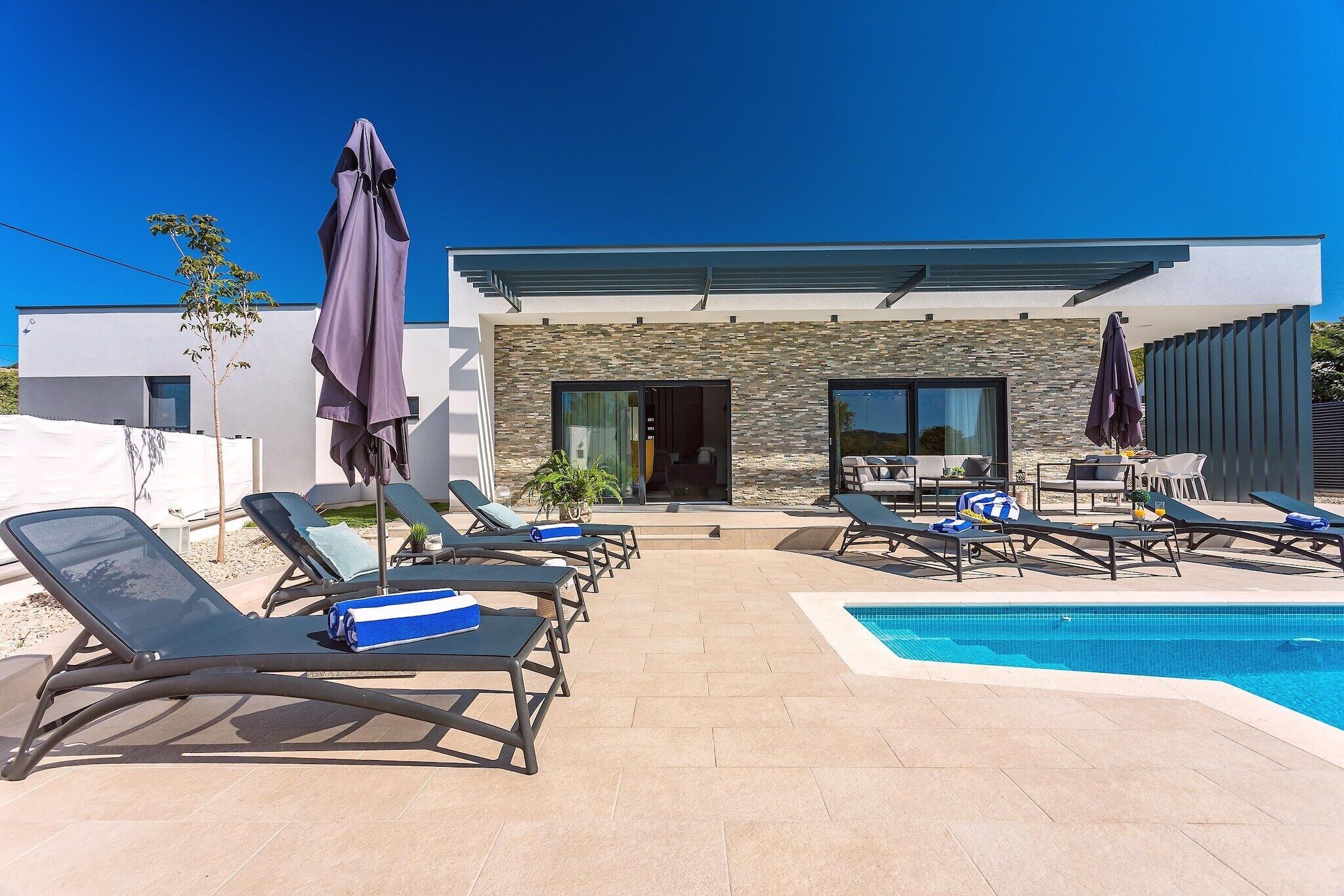 A poolside area with lounge chairs and umbrellas, a modern building with stone facade, and a clear blue sky.