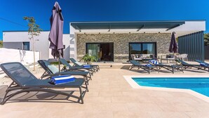 A poolside area with lounge chairs and umbrellas, a modern building with stone facade, and a clear blue sky. - Luxury Villa Retreat in Šestanovac (Šestanovac)