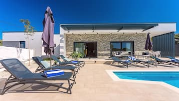 A poolside area with lounge chairs and umbrellas, a modern building with stone facade, and a clear blue sky.