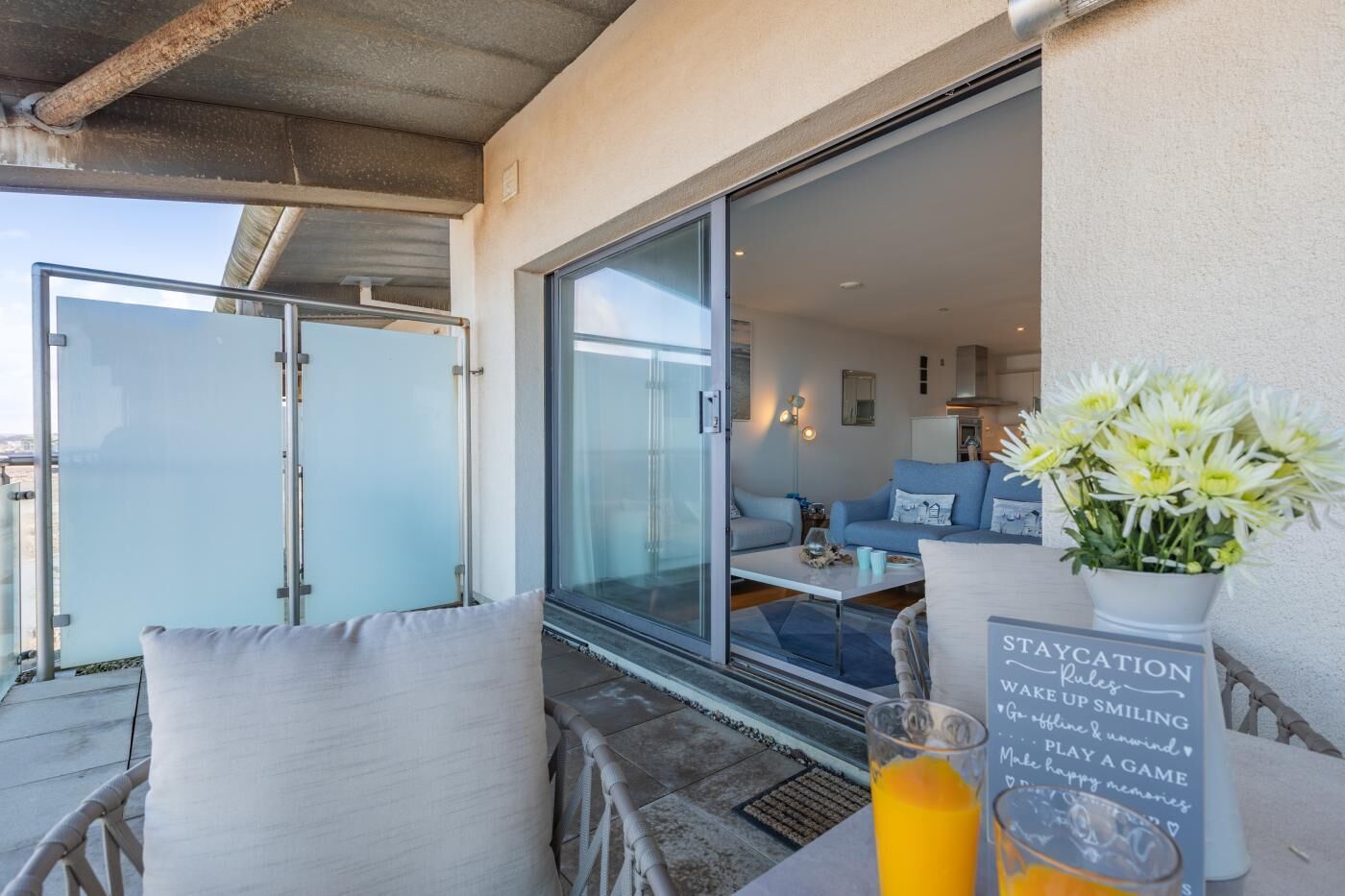 A balcony with a glass door leading to a living area, a table with a glass of orange juice, and a sign with staycation rules.