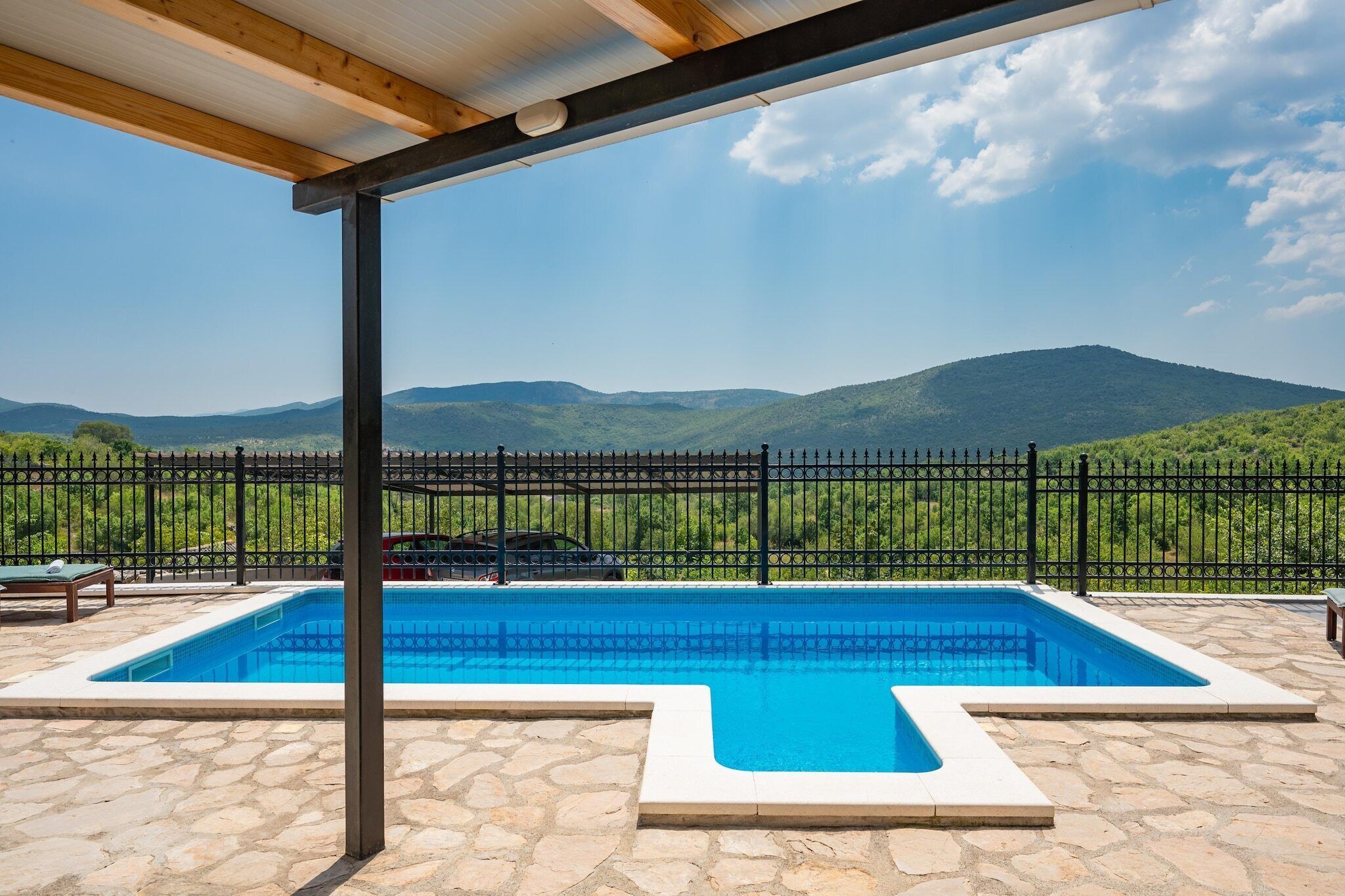 Una piscina con un patio de piedra, una pérgola de madera y vistas a colinas verdes.