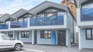 Een modern meerlaags gebouw met grote ramen en balkons, een surfboard tegen de muur en een geparkeerde auto ervoor.