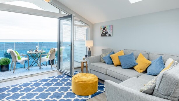 Een moderne woonkamer met een grijze bank, gele en blauwe kussens, een geel poufje en een blauw kamerkleed. Een glazen deur leidt naar een balkon met uitzicht op zee.