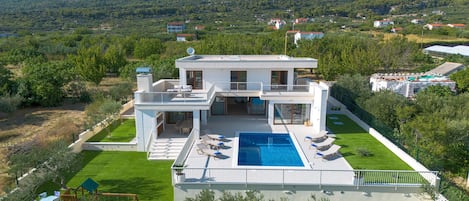 A modern two-story house with a swimming pool, surrounded by greenery and hills.