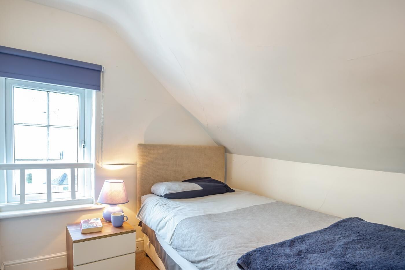 Ein kleines, gemütliches Schlafzimmer mit einem Einzelbett, einem Nachttisch, einer Lampe und einem Fenster mit Blick hinaus.