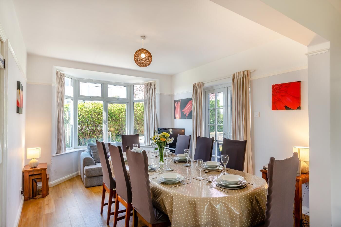 A dining room with a round table set for six, large windows, and a view of greenery outside.