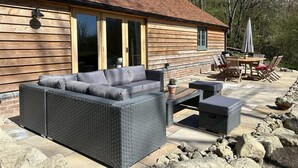 Une maison en bois dotée d’un patio comprenant un canapé en osier gris, une table avec des chaises et un pot de fleurs.