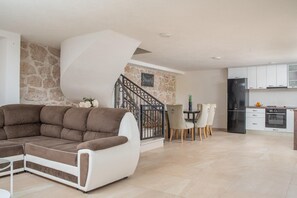 Ein modernes Wohnzimmer mit einer Eckcouch, einem Essbereich mit Stühlen und einem Tisch, einer Küche mit weißen Schränken und Geräten sowie einer Wand mit Steinverkleidung.