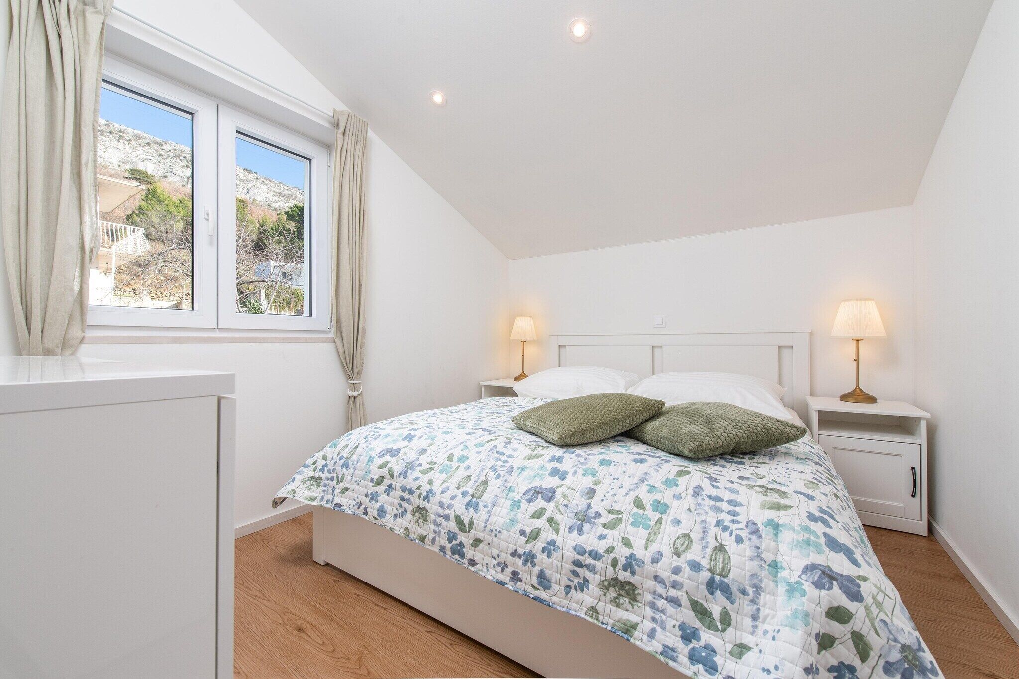 Um quarto com cama, duas mesas de cabeceira com luminárias, uma janela com cortinas e vista para o exterior.