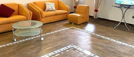 Una sala de estar con un conjunto de sofás, una mesa de centro de vidrio, una alfombra estampada y un televisor.