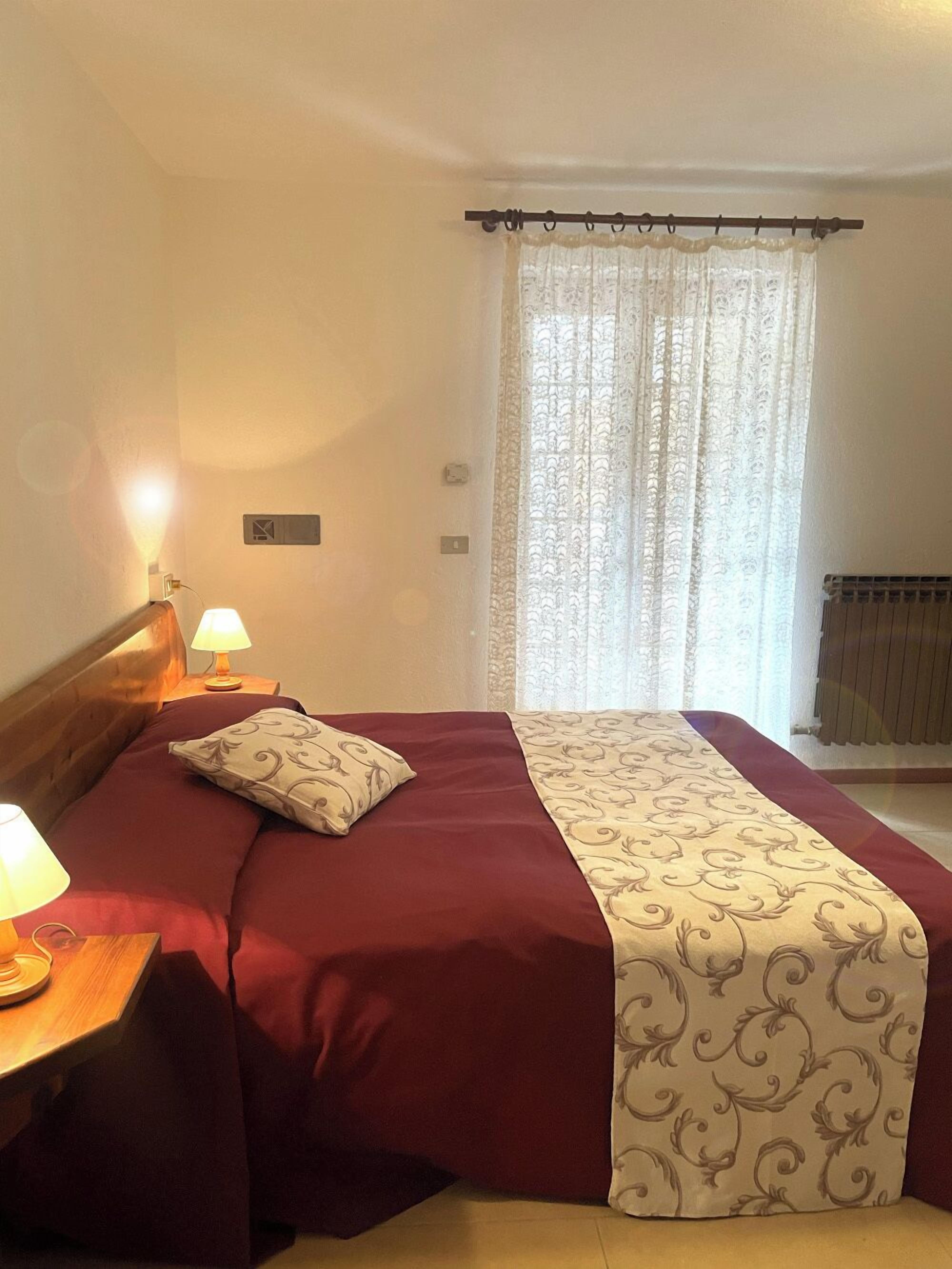 Una cama bien hecha con una colcha roja y blanca, dos lámparas de noche y una ventana con cortina.