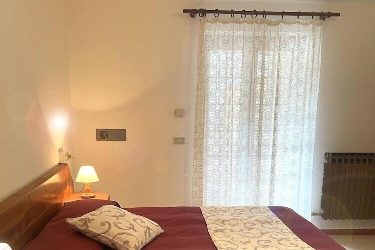 A hotel room with a bed, two bedside lamps, a window with a curtain, and a radiator.