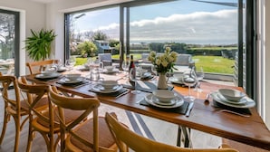 Un coin repas avec une table dressée pour six personnes, de grandes fenêtres donnant sur l’extérieur et une plante en pot placée à gauche.