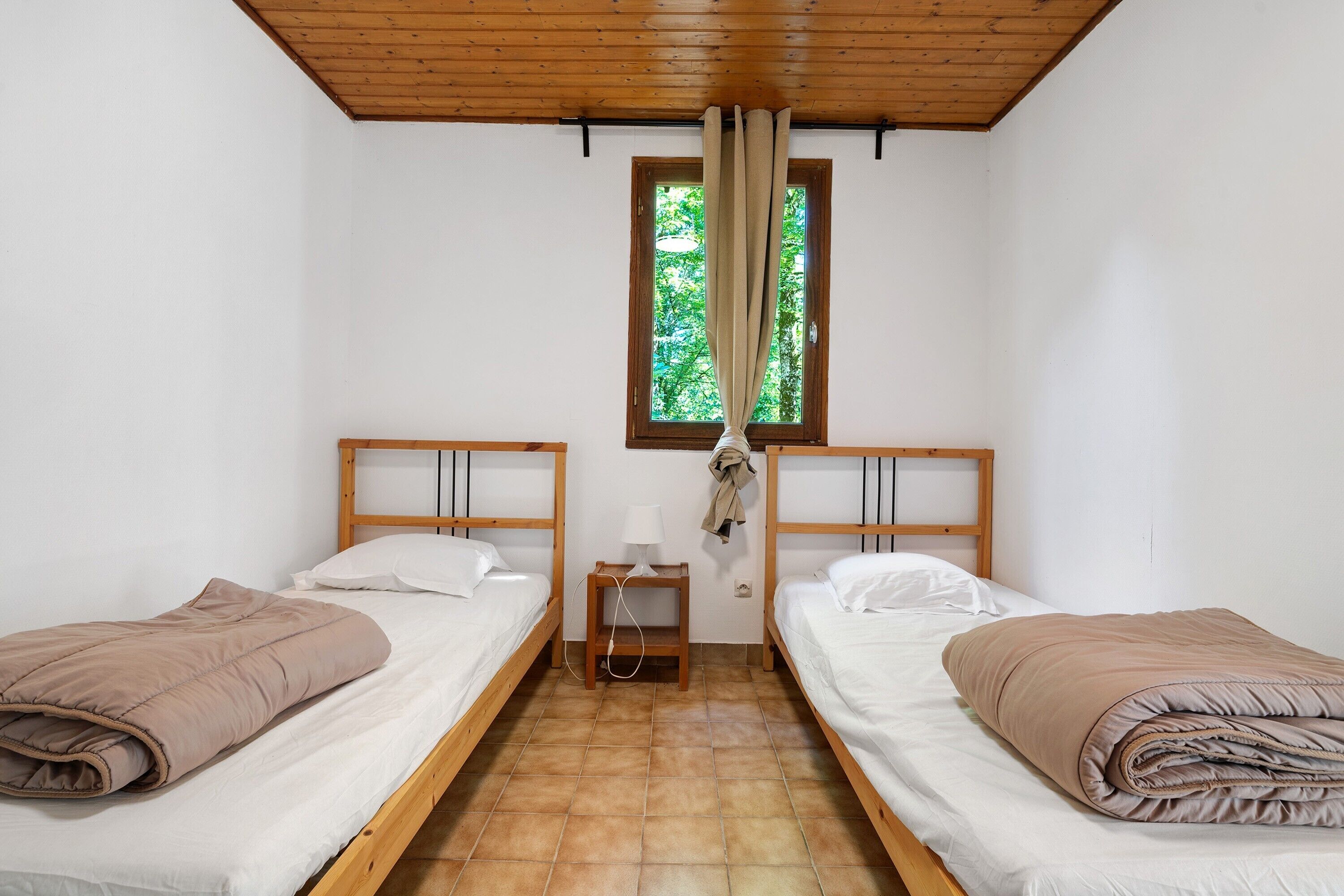 A room with two single beds, a wooden ceiling, a window with curtains, a nightstand with a lamp, and a wall socket.