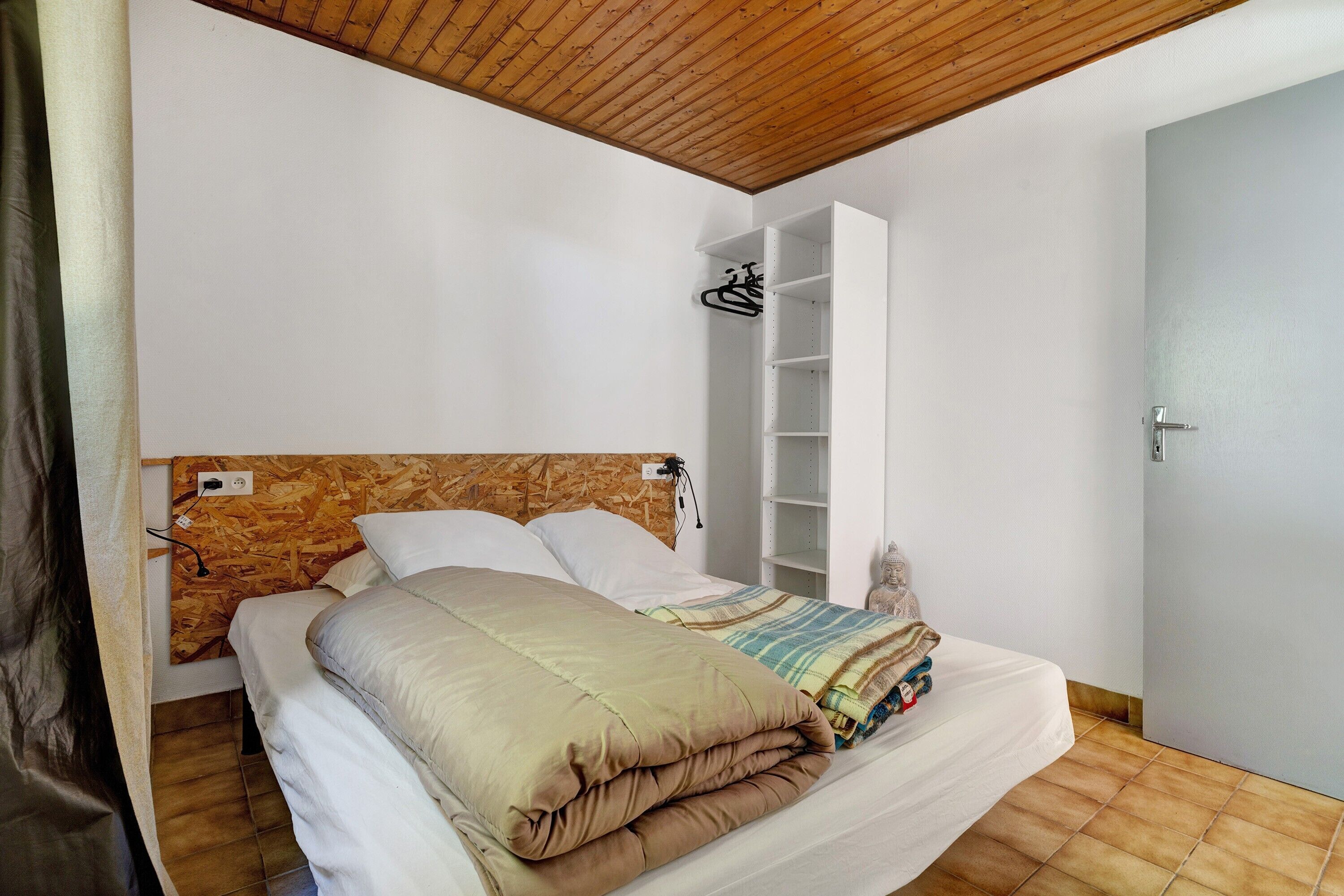 A bedroom with a wooden headboard, a bed with a beige and white duvet, a white wardrobe, and a tiled floor.
