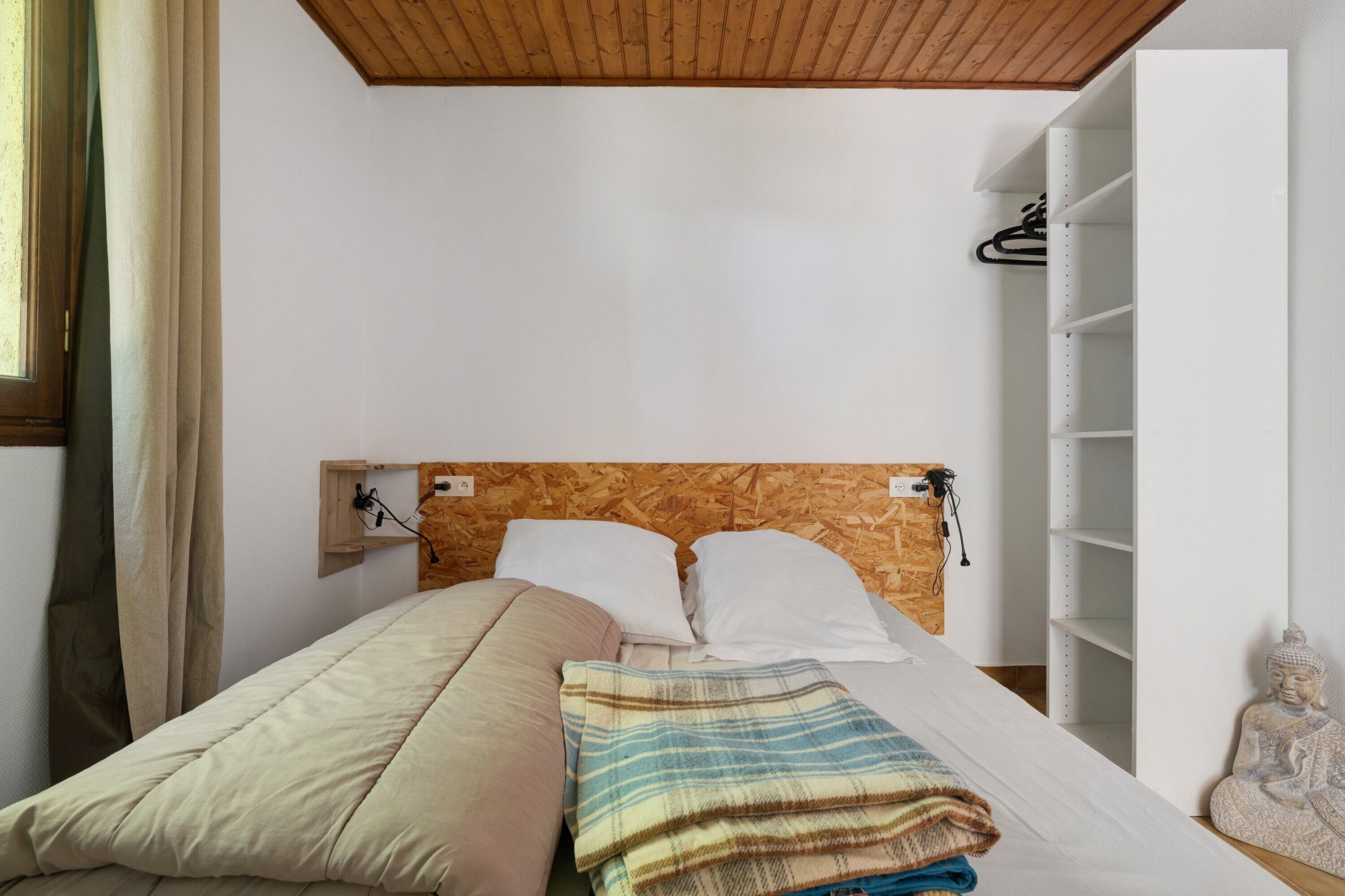 A bed with a wooden headboard, a white wall, and a shelf with a statue.
