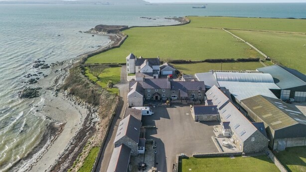 Vue aérienne d’une propriété située sur la côte, avec un grand bâtiment, un champ herbeux et une plage.