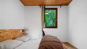 Quarto com cabeceira de madeira, janela com vista para árvores, cama com roupa de cama branca e castanha e um abajur.