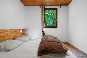 Une chambre à coucher avec une tête de lit en bois, une fenêtre donnant sur des arbres, un lit avec une literie blanche et marron, et une lampe de chevet.