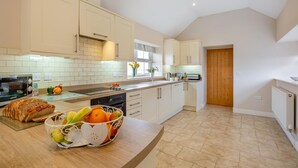 A modern kitchen with white cabinets, a large island with fruits, a toaster, and a window with flowers. - 3 Bed in Llanfwrog (oc-f31053) (Llanfaethlu)