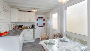 Eine Küche mit weißen Schränken, ein Essbereich mit Tisch für zwei Personen und ein Blick durch ein Fenster.