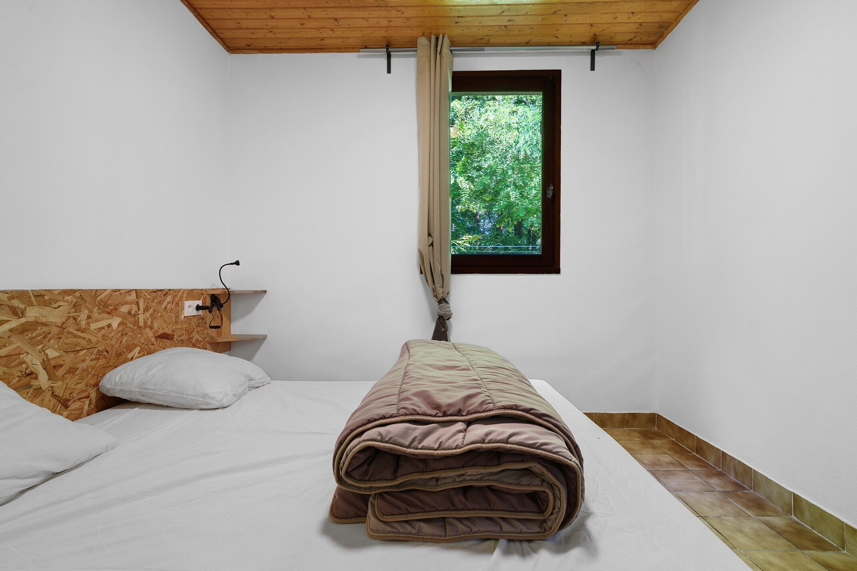 Una camera piccola con un letto singolo, una testiera in legno, una finestra con vista su piante e un soffitto in legno.