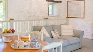 A cozy living room with a light blue sofa, a wooden dining table with a fruit bowl and a cake, and a white railing. - 2 Bed in Launceston (oc-d31000) (Trevadlock)
