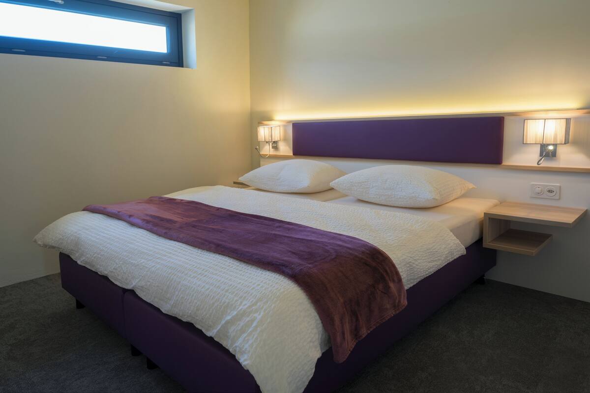 Une chambre d’hôtel moderne avec un grand lit, deux tables de chevet avec des lampes, une fenêtre et une étagère intégrée.